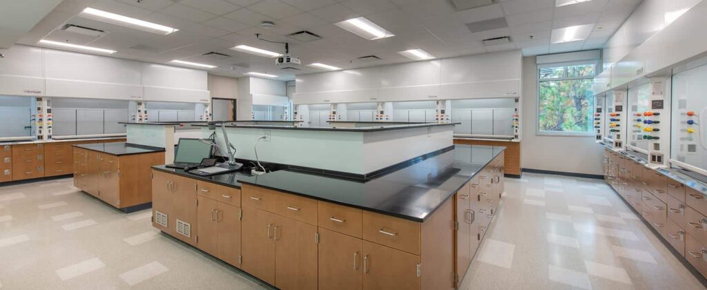 A modern science laboratory featuring black countertops, wooden cabinets, a laptop, safety equipment, and bright overhead lighting. Large windows allow natural light to fill the clean and empty room.