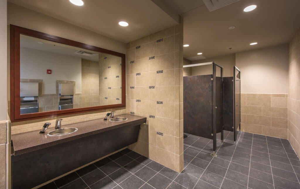 A tidy public restroom featuring two sinks beneath a large mirror, with light brown tiled walls and a black-tiled floor. In the background, two stalls with partially open dark doors are visible.