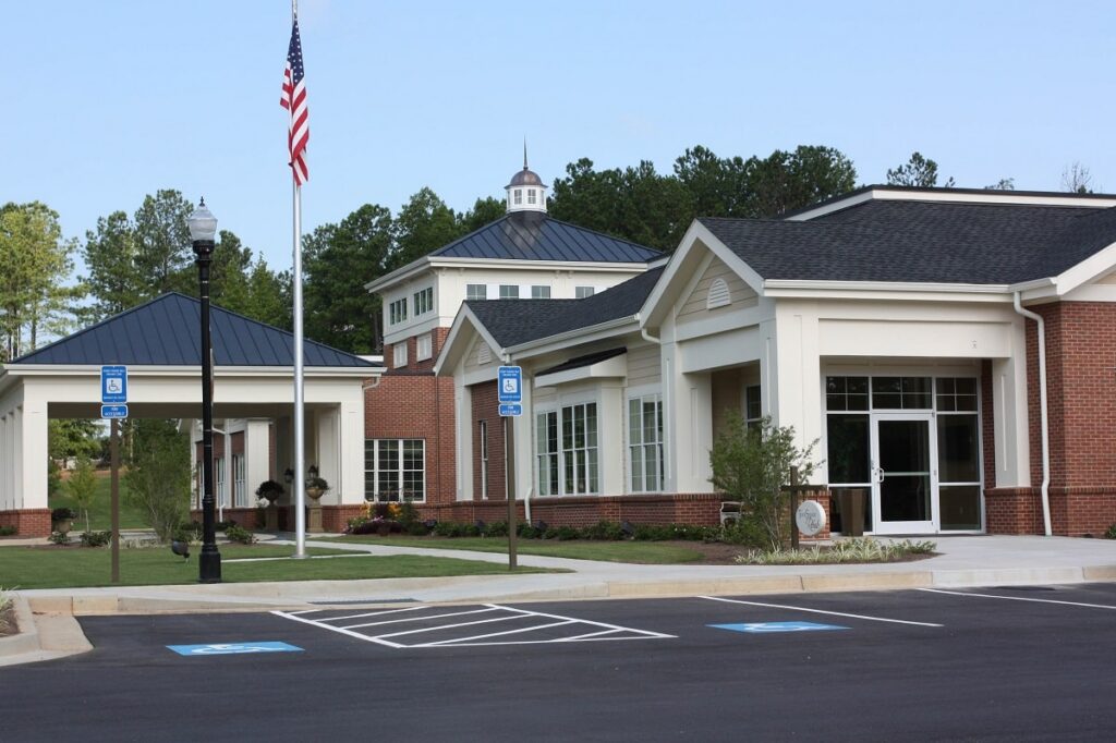 A brick building with white trim, an American flag, designated handicap parking spaces, and a small tower, surrounded by trees and greenery beneath a clear blue sky.