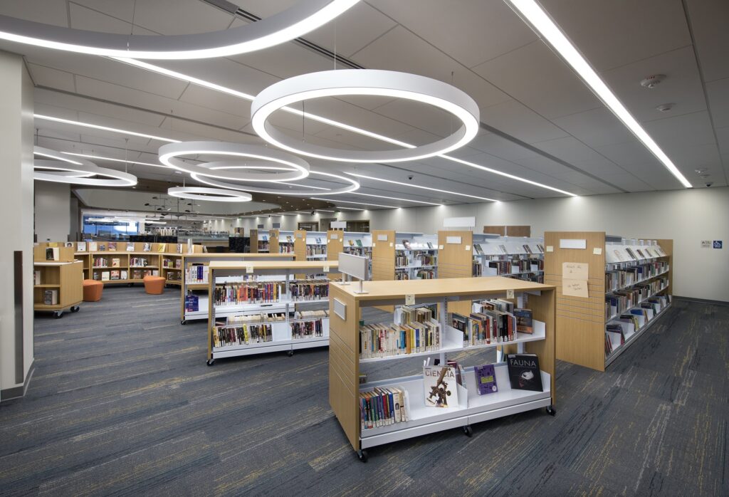 Modern library interior with wooden bookshelves filled with books, circular ceiling lights, and a carpeted floor. Some orange chairs and large windows are visible in the background, creating a bright and open atmosphere.