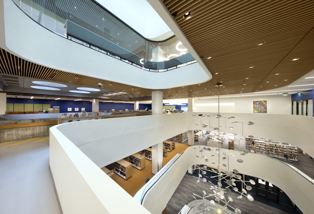 A modern library interior featuring curved white balconies, wooden ceiling panels, and rows of bookshelves. The space is open and airy, with natural light streaming through skylights and a central light fixture adding to the inviting atmosphere.