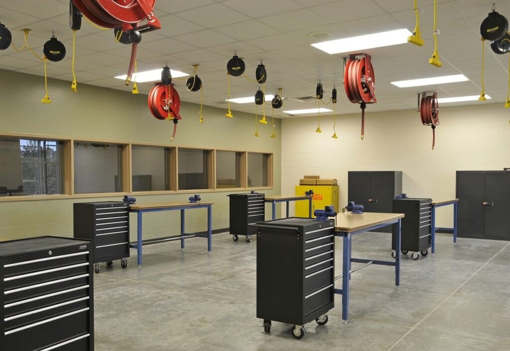 A workshop room featuring multiple workbenches, mobile tool cabinets, wall-mounted cabinets, red hose reels and yellow drop cords suspended from the ceiling, and windows lining one wall.