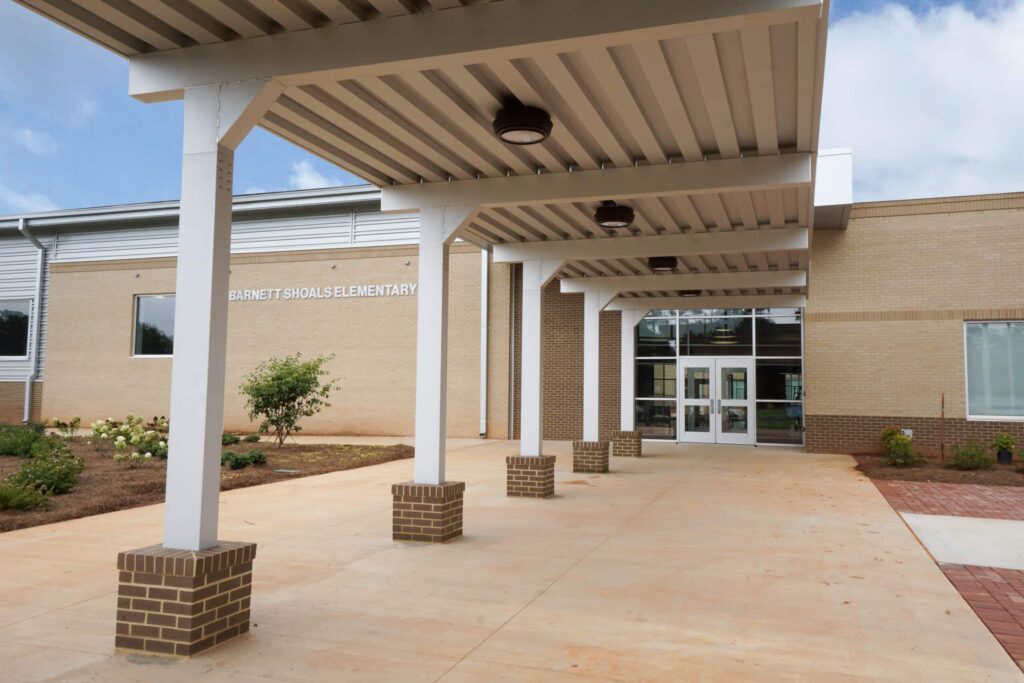 A covered walkway leads to the entrance of Barnett Shoals Elementary, a modern brick and glass school building, on a bright day.