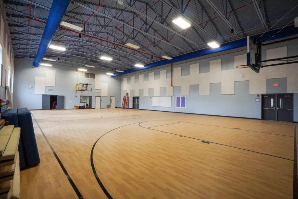 An empty indoor gymnasium featuring a light wood floor, basketball hoops, a high ceiling, and fluorescent lighting. The gray walls have white panels, with doors and windows lining the sides.