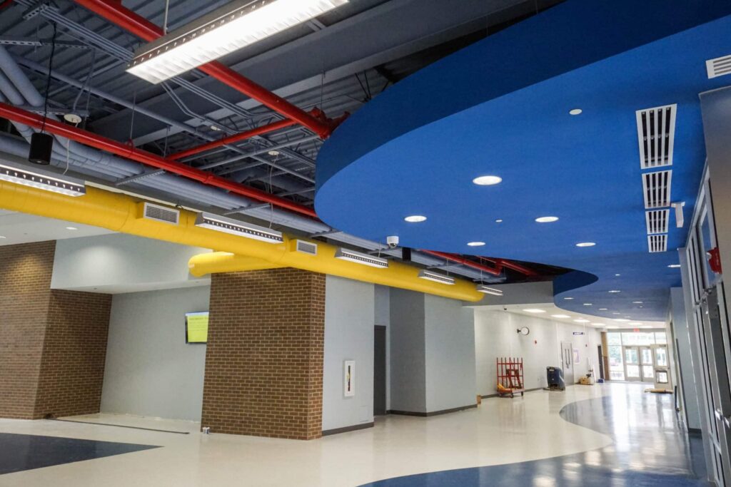 A contemporary hallway features a blue ceiling, exposed red pipes, yellow vents, and white walls with brick accents. Overhead lights illuminate the shiny floor, which displays blue and cream hues.