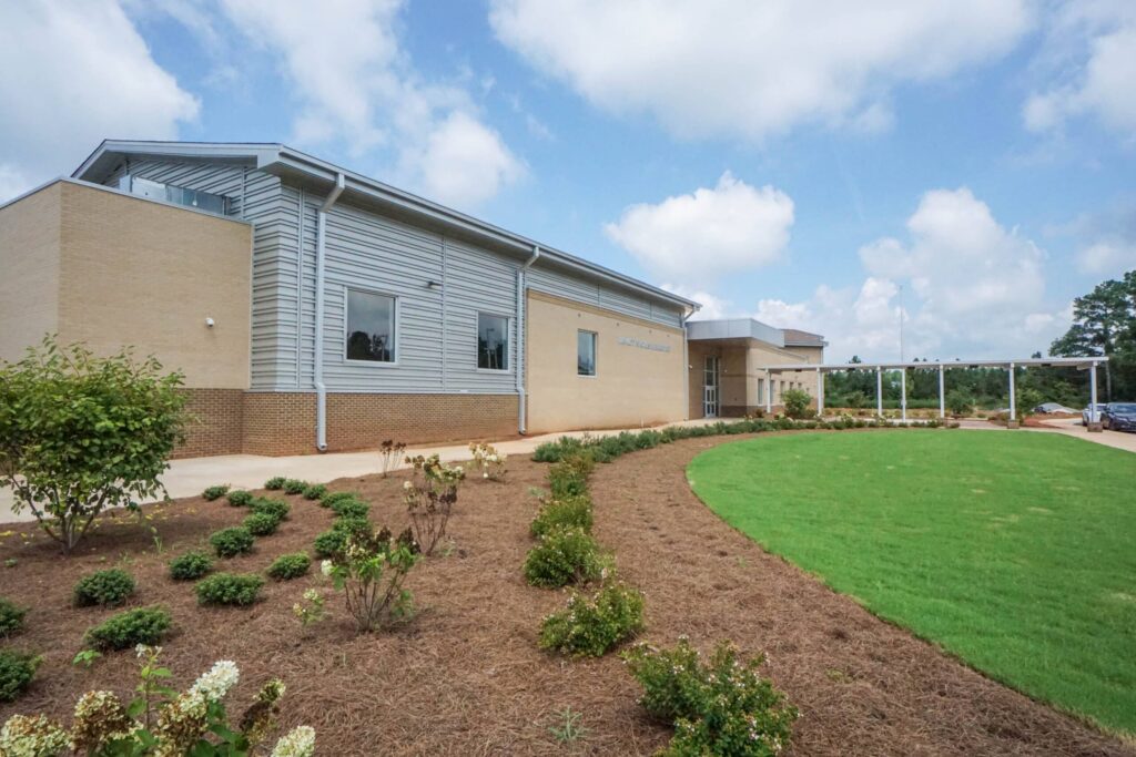 A contemporary single-story building featuring tan brick and metal siding, bordered by neatly landscaped bushes, mulch beds, and a vibrant green lawn beneath a partly cloudy sky.