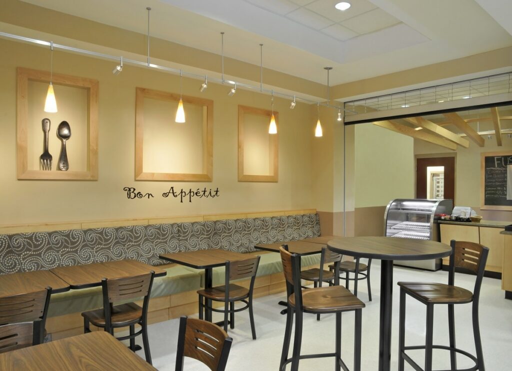 A modern cafe with wooden tables and chairs, cushioned bench seating, framed art, pendant lights, and Bon Appétit written on the wall; an empty display case is visible in the back.