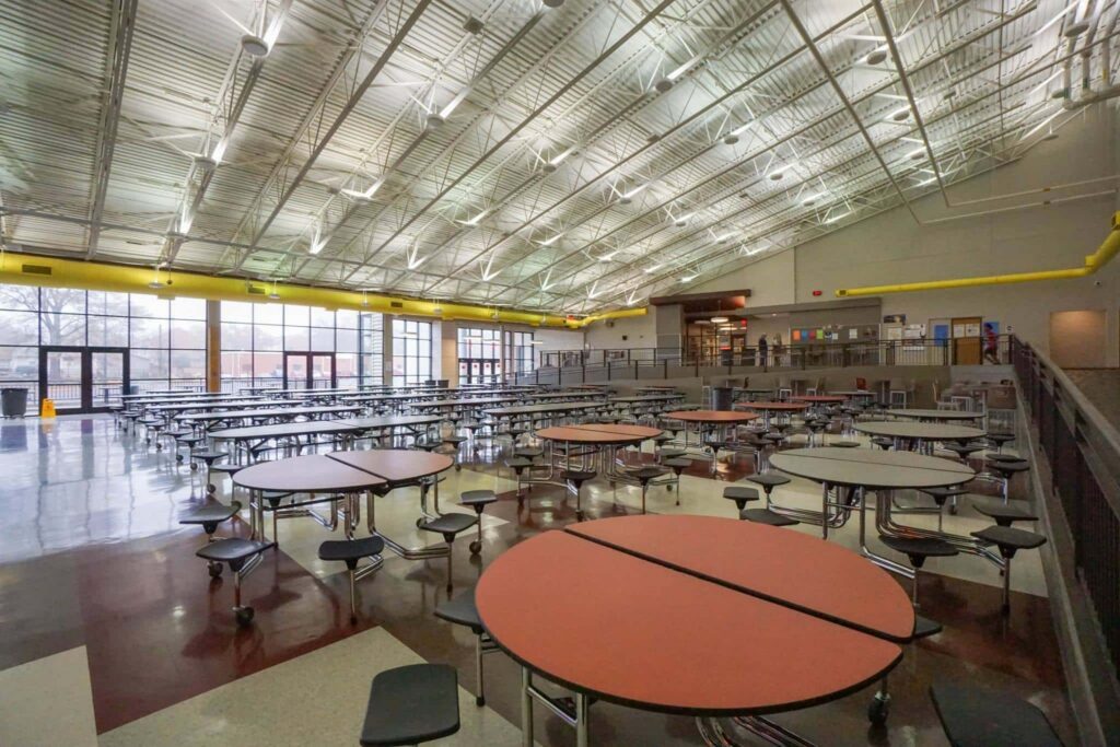 Large, empty cafeteria featuring rows of round tables with attached stools, shiny floors, a high ceiling with exposed beams, and expansive windows allowing in natural light. The space is unoccupied.