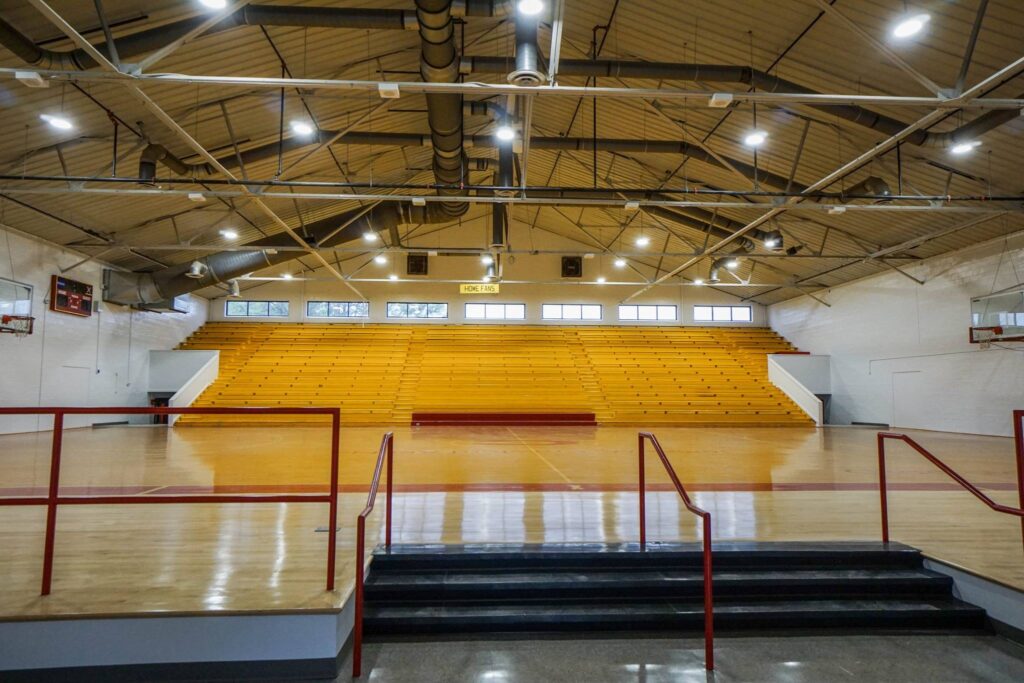 Empty indoor gymnasium featuring wooden bleachers, a shiny basketball court, red railings, and bright overhead lights. The area is spotless and tidy, with no one visible.