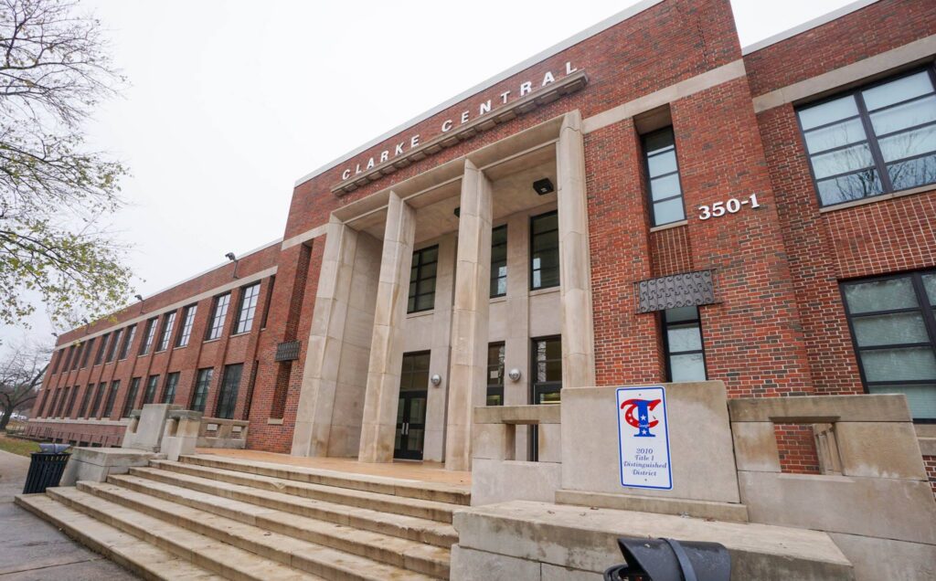 A brick building with tall columns at the entrance, labeled Clarke Central. Steps lead to glass doors. A sign with the school’s logo and address, 350-1, is visible near the front. Trees line the side.