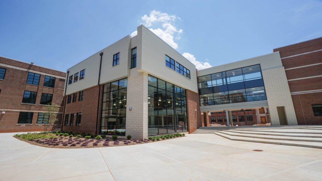 Modern school building featuring large windows, red brick and white accents, landscaped greenery on the left side, and wide concrete steps that lead to the entrance beneath a bright blue sky with a few clouds.