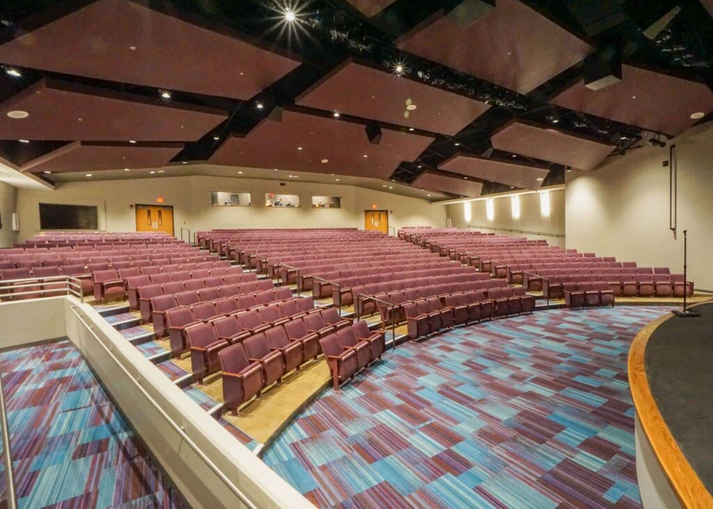 An empty auditorium featuring rows of purple seats, a blue and purple patterned carpet, and a spacious stage on the right. The ceiling shows both overhead lights and acoustic panels.