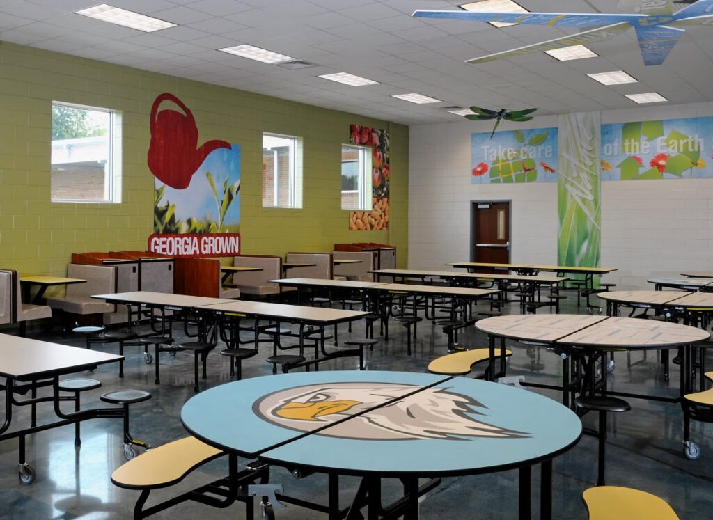 A school cafeteria featuring a mix of round and rectangular tables with yellow seats, murals of nature scenes on the walls, and a prominent eagle graphic displayed on one table. The space is well-lit by high windows that let in plenty of daylight.