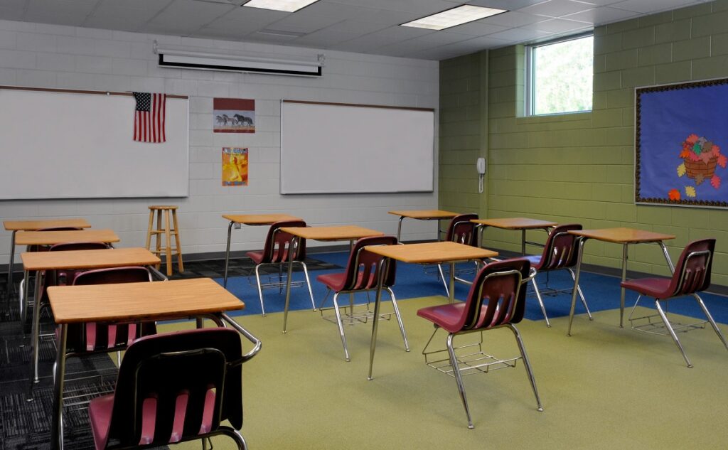 Empty classroom featuring wooden desks and maroon chairs, whiteboards mounted on the wall, an American flag, a stool, various posters, a window, green-painted walls, and a colorful bulletin board on the right side.