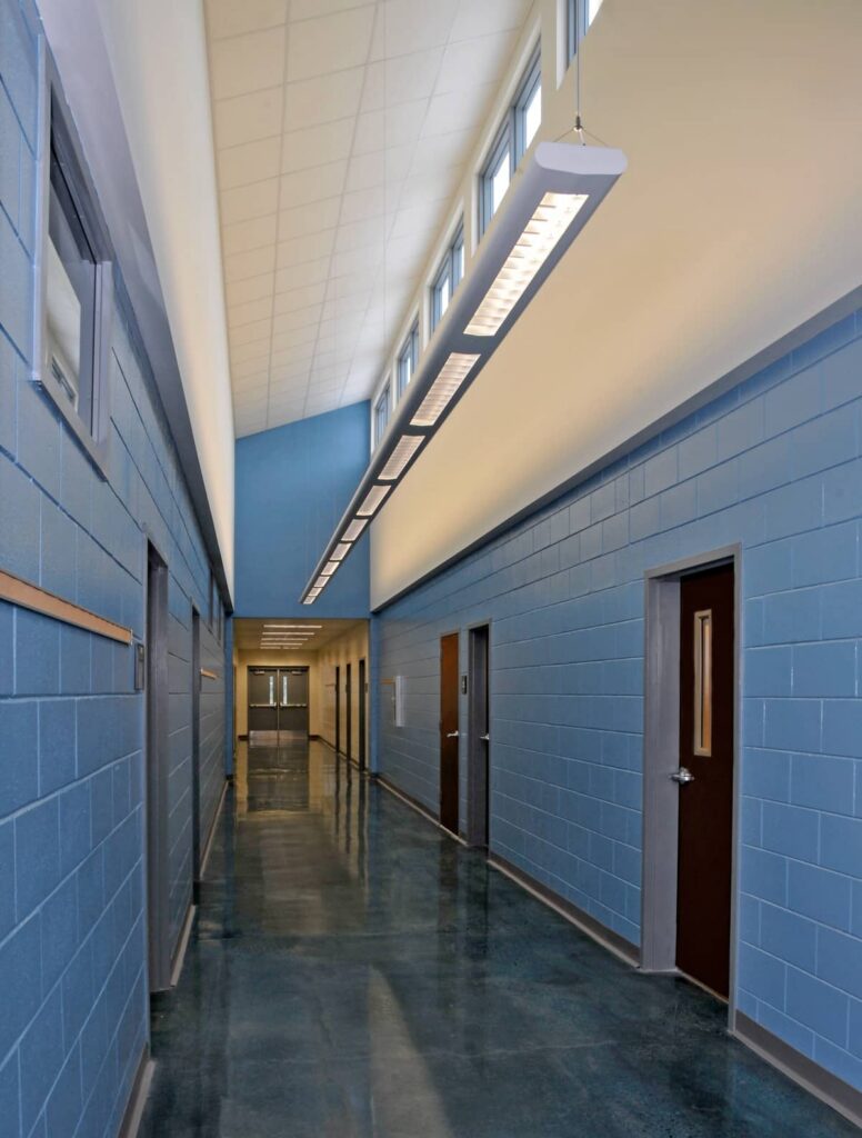 A hallway featuring blue walls, multiple brown doors with small windows, a shiny dark floor, and a tall ceiling fitted with large light fixtures and upper windows that allow natural light to stream in.
