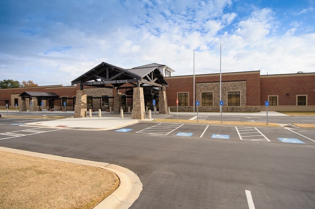 A single-story brick and stone school building with a covered entrance, surrounded by an empty parking lot featuring several accessible parking spaces. The partly cloudy sky is visible above.