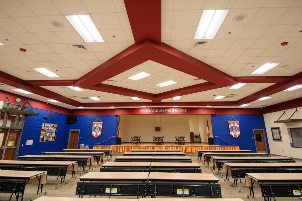 A spacious, empty cafeteria or assembly hall features rows of tables and chairs arranged toward a stage. The walls are painted blue and red, with banners hanging on both sides of the stage.