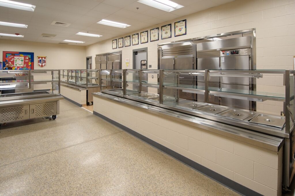 An empty stainless steel cafeteria serving line in a spotless, well-lit room, featuring trays and glass sneeze guards. The space has beige walls adorned with framed certificates and a colorful wall poster in the background.