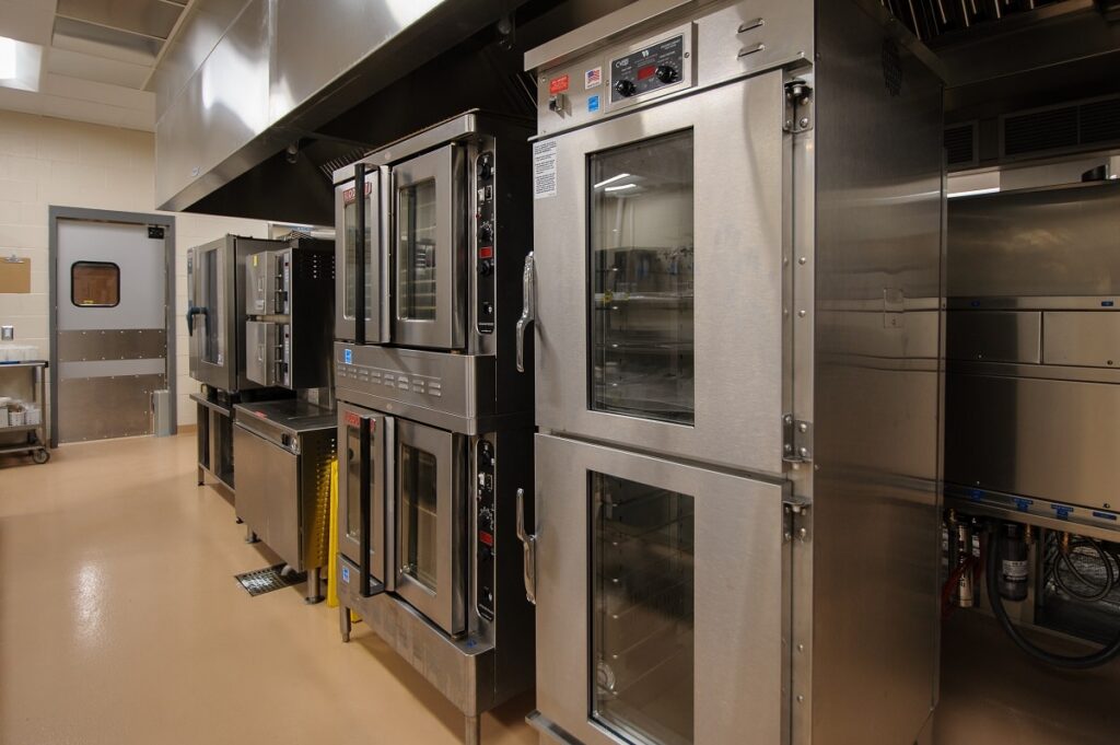 Commercial kitchen featuring stainless steel double-door ovens, metal counters, and assorted equipment. The space has beige floors, white walls, and fluorescent ceiling lights. A gray door can be seen in the background.