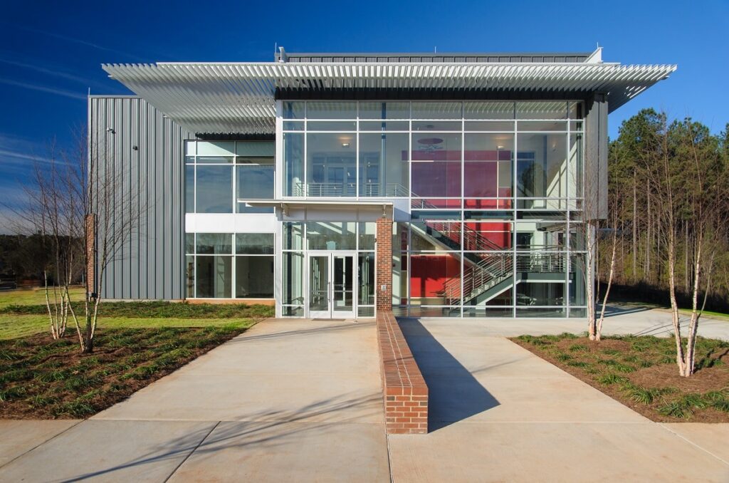 Modern two-story building with large glass windows, metal siding, and a prominent overhang, surrounded by landscaped greenery and a clear blue sky. A brick walkway leads to the main entrance.