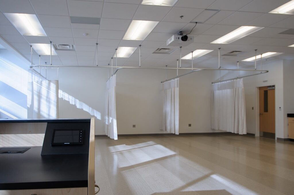 A brightly lit, empty hospital room featuring several curtained patient areas, a ceiling-mounted projector, and a monitor positioned on a counter in the foreground. Sunlight pours through windows along the left wall.