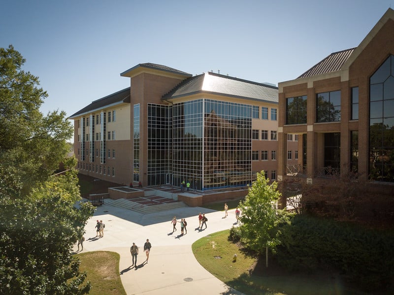 A contemporary university building with expansive windows stands in sunlight as students walk along nearby pathways. Trees around the building create shaded patterns on the ground.