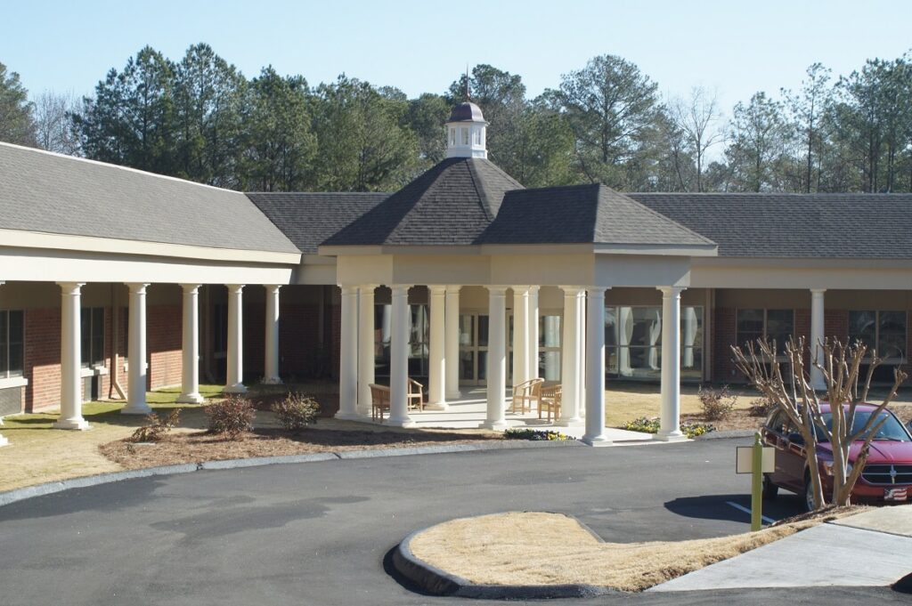 A building featuring white columns and a covered entrance stands before a curved driveway, surrounded by trees and with a red car nearby beneath a clear sky.