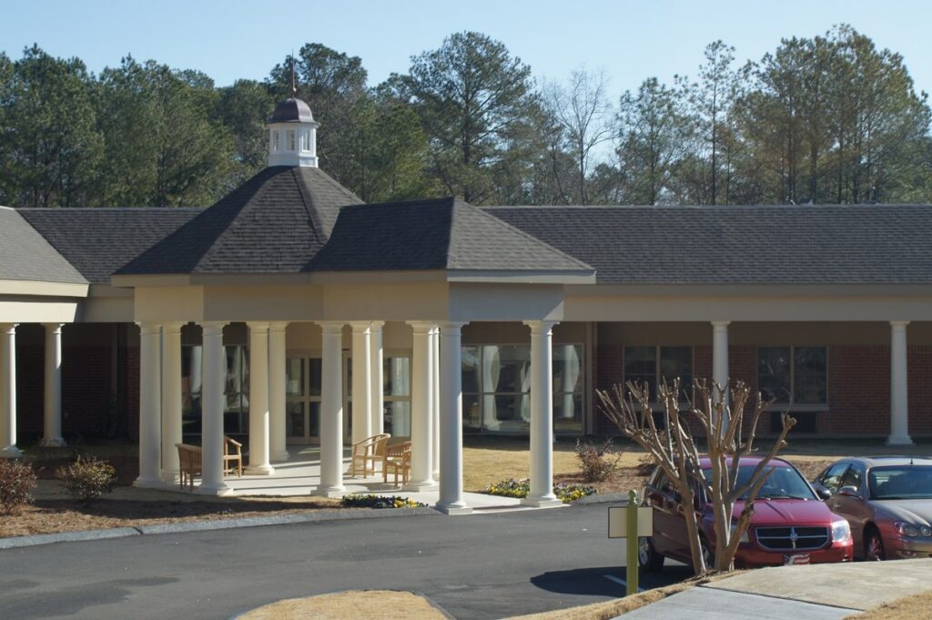 A building with a covered entrance supported by white columns, featuring a small cupola on top. Two wooden benches are under the canopy, and cars are parked in front. Trees are visible in the background.