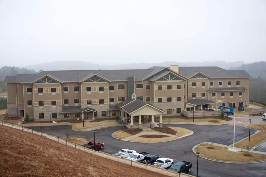 A large three-story beige building with multiple entrances and a parking lot in front, set amid grassy areas and rolling hills beneath a cloudy sky. Several cars are parked outside.