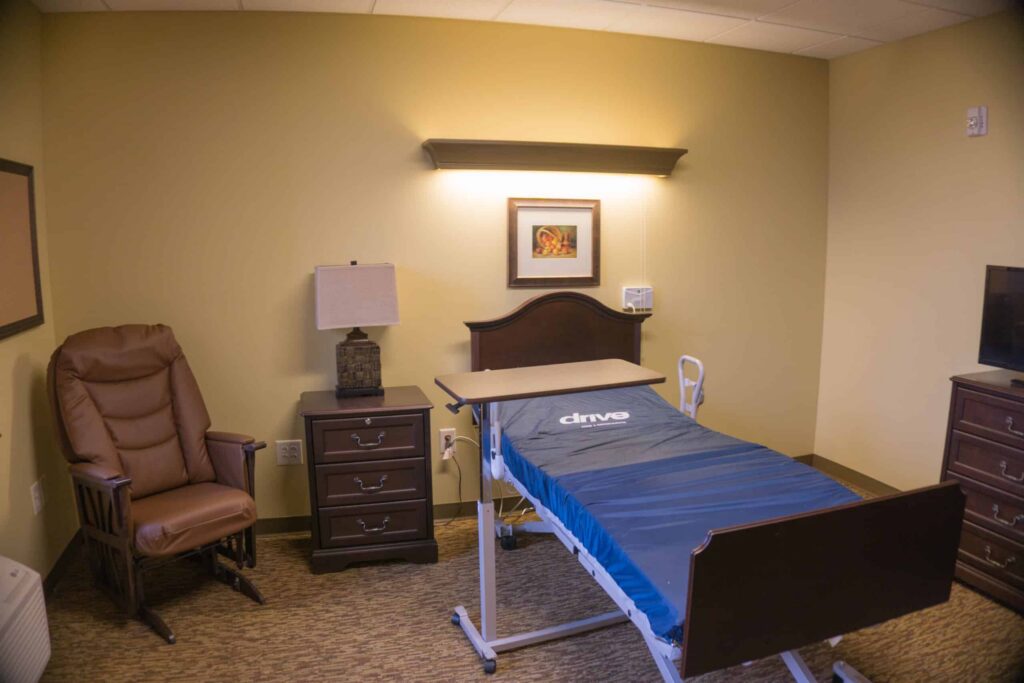 A neatly arranged room with a single medical bed, a brown recliner, a nightstand with a lamp, a dresser, and a wall picture. The walls are painted yellow and the room has warm lighting.