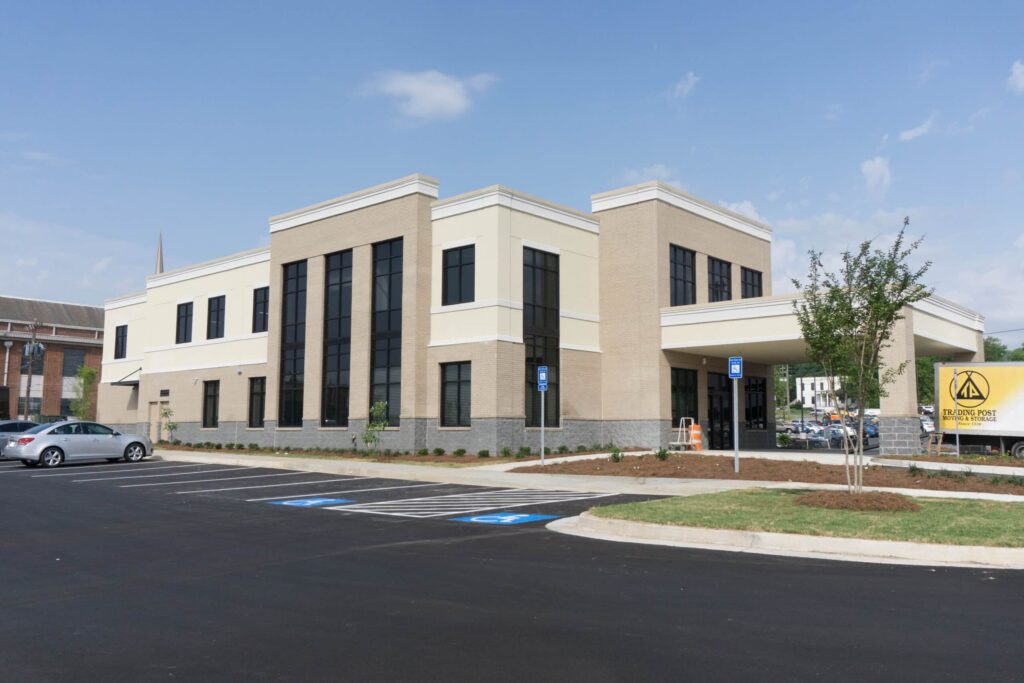 A modern two-story beige and cream commercial building with large windows, a small parking lot with accessible spaces, and a sign reading Elmore Family. Trees and cars are visible around the building.