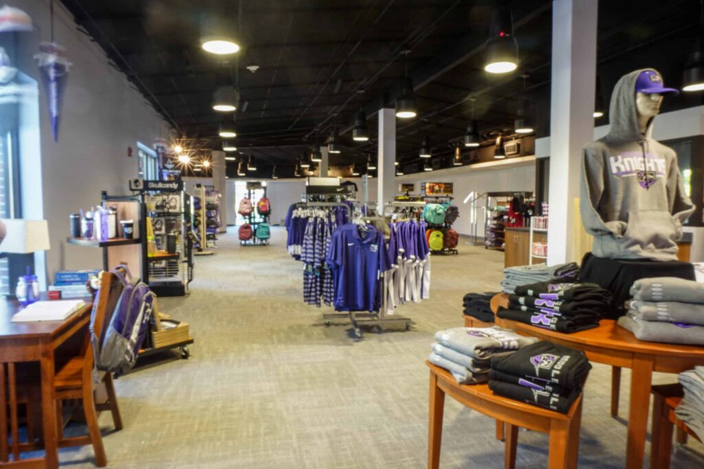 Campus bookstore interior featuring racks of clothing, school merchandise, and a mannequin dressed in a hoodie and cap. Tables and shelves display neatly folded shirts and accessories.