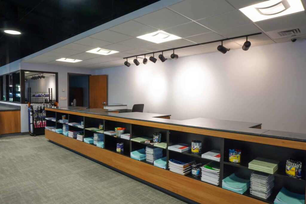 A modern office supply counter with shelves holding notebooks, clipboards, paper, and pens. The countertop is empty, and the background wall is plain and well-lit. A chair and open door are visible behind the counter.