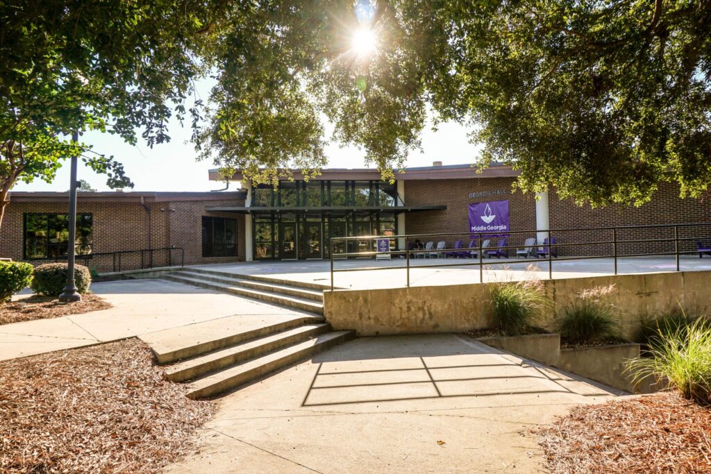 Sunlight filters through tree branches, illuminating a brick building that has large glass windows, a ramp, and steps leading up to its entrance. Near the doors, purple banners are displayed. The walkway is bordered by shrubs and mulch.