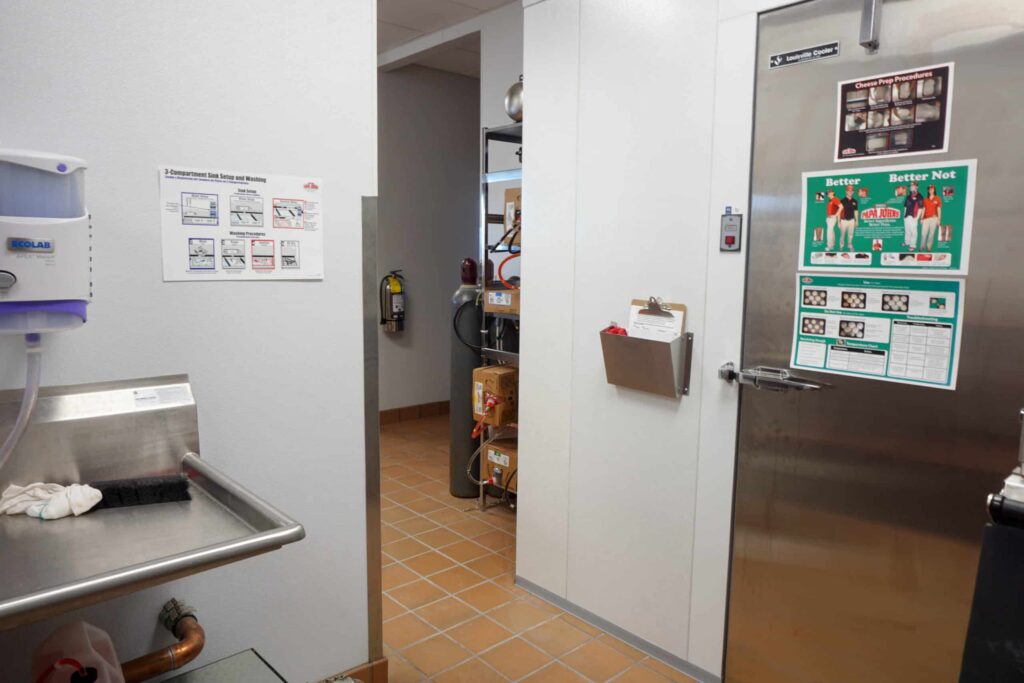 A clean commercial kitchen featuring a metal sink, a walk-in refrigerator, safety posters displayed on the wall, shelves stocked with boxes, and a fire extinguisher visible through an open doorway.