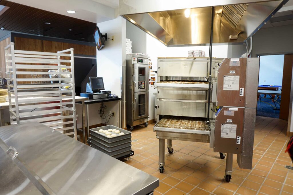 Commercial kitchen featuring stainless steel counters, an industrial oven with trays, a computer station, baking racks, and trays of dough balls on tiled flooring. Through an open door, a room with a ping pong table can be seen.