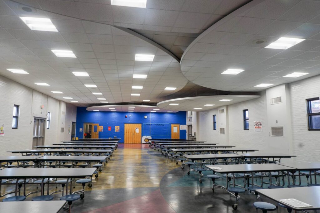 A vacant school cafeteria featuring rows of gray tables and benches, a shiny multicolored floor, bright overhead lights, and a blue wall at the back with several doors and a clock.