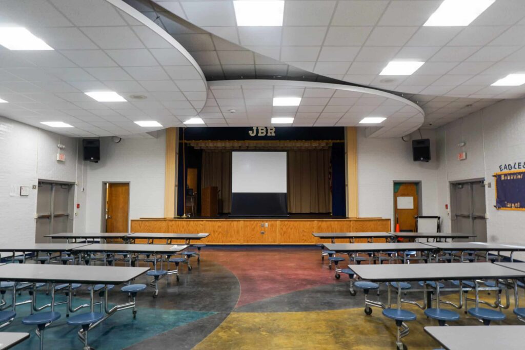 A school cafeteria with gray tables and benches, colorful flooring, and a stage at the front with a projector screen and the letters JBR above it. Banners hang on the walls.