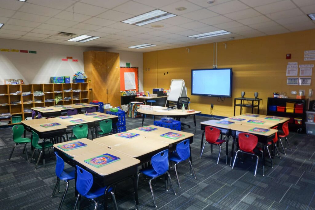 A brightly lit classroom featuring grouped desks, vibrant chairs, shelves stocked with bins, a whiteboard, and a large screen at the front. Educational posters and various supplies are arranged throughout the room.