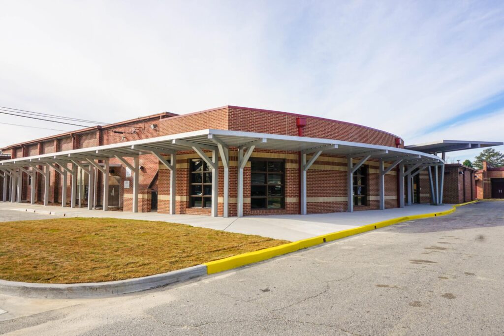 A contemporary brick building featuring expansive windows and a sleek metal canopy, outlined by a yellow curb. It sits beside a driveway and a patch of grass, all beneath a sky with scattered clouds.