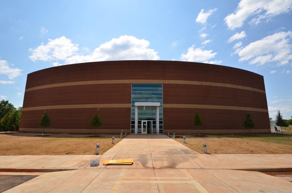 A spacious, contemporary brick building with curved architecture and a glass front entrance, bordered by young trees beneath a blue sky dotted with scattered clouds.