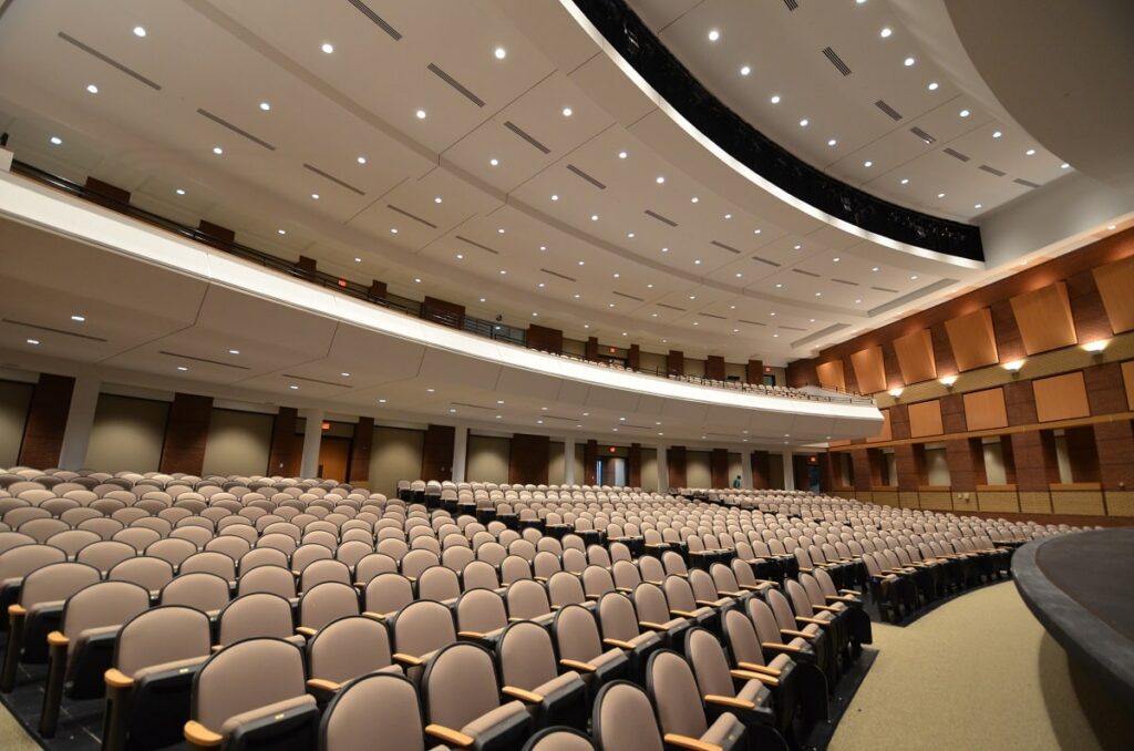Large modern auditorium featuring rows of empty beige seats, a curved balcony, and bright ceiling lights, all oriented toward a wide stage.