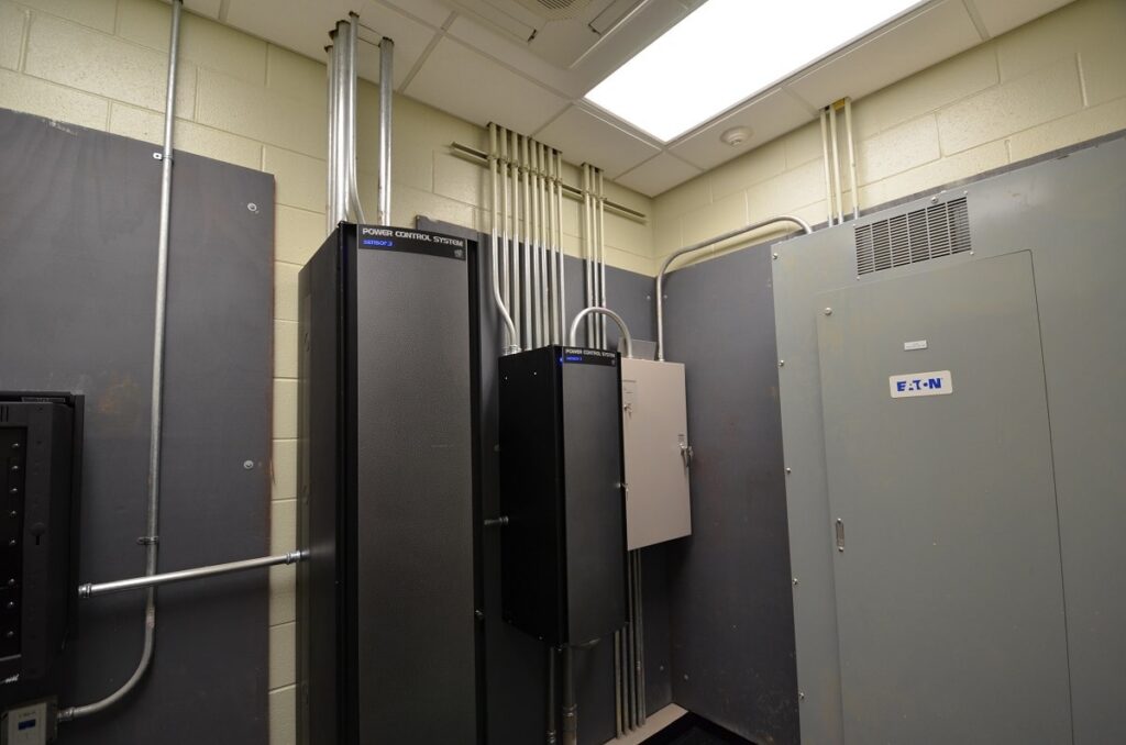 Small utility room with electrical panels, conduit pipes running to the ceiling, and a large gray cabinet labeled EATON on the right wall under bright fluorescent lighting.