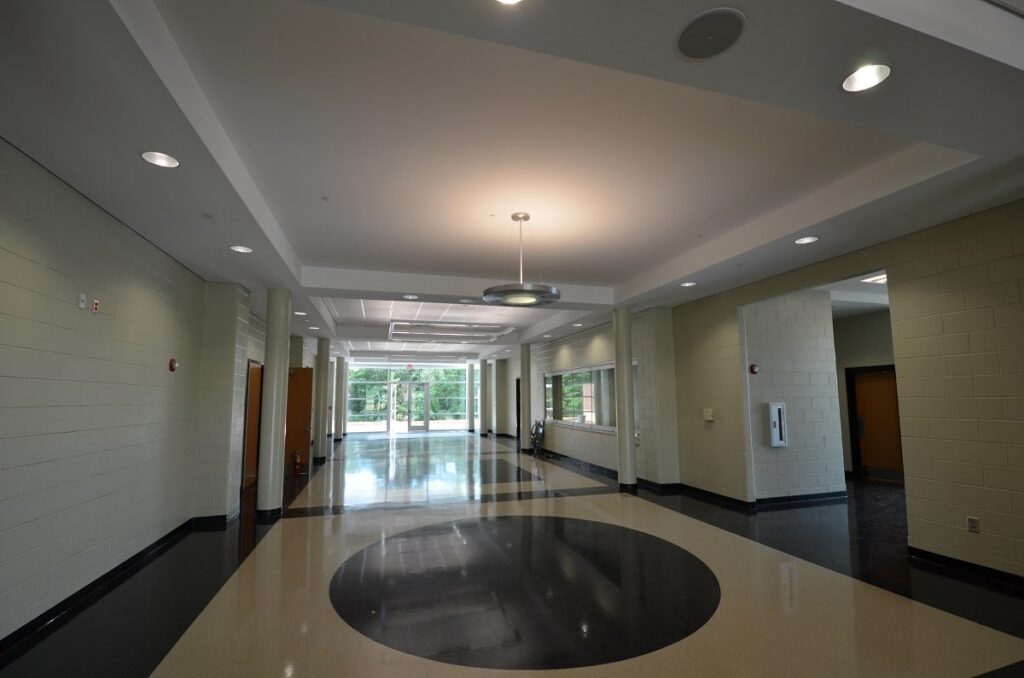 A modern hallway featuring cream and black tiled flooring, overhead lights, cream brick walls, windows, and glass double doors that open to a lush, tree-filled outdoor area.