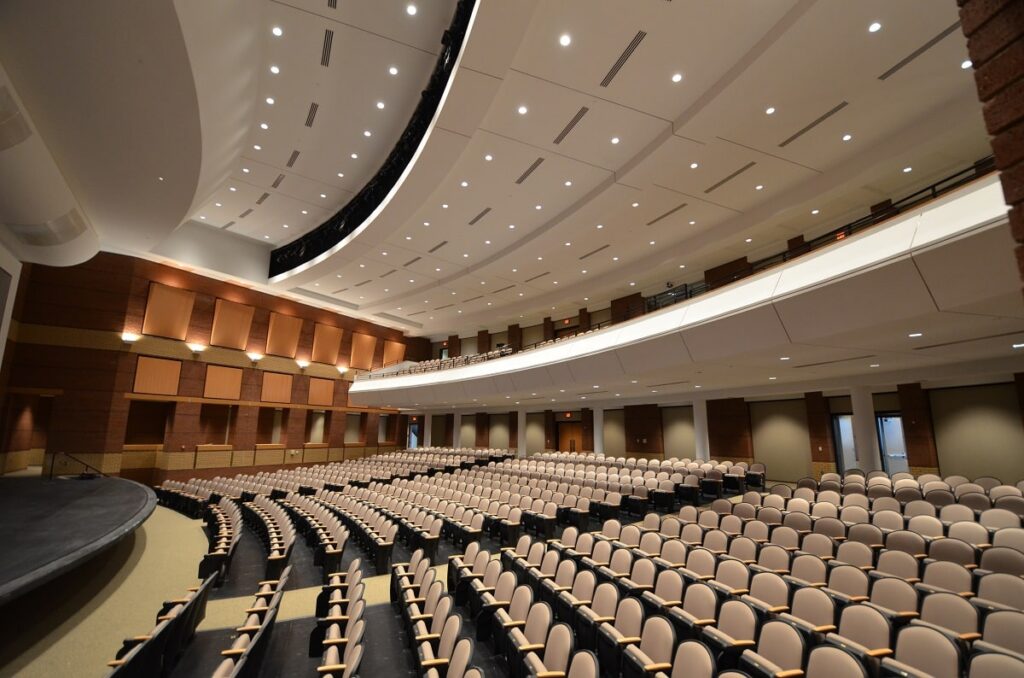 A spacious modern auditorium featuring rows of empty beige seats, curved balconies, and a large stage on the left. The ceiling is equipped with recessed lighting and acoustic panels.