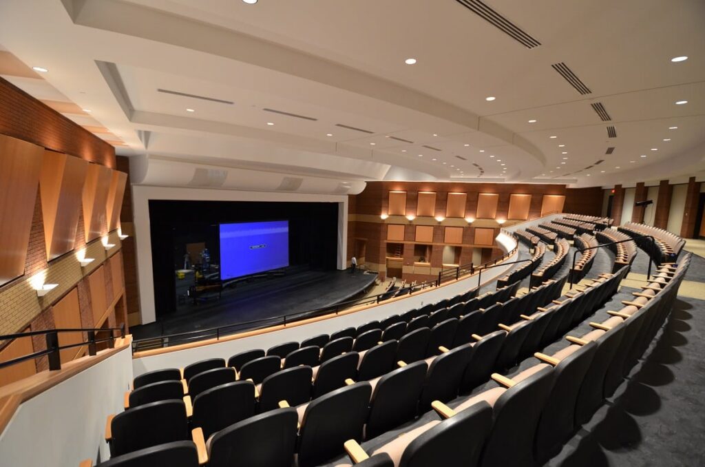 Empty modern auditorium with curved rows of black seats, wooden accents, and a stage featuring a blue projection screen. The space is well-lit, with overhead lights and wall sconces illuminating the interior.