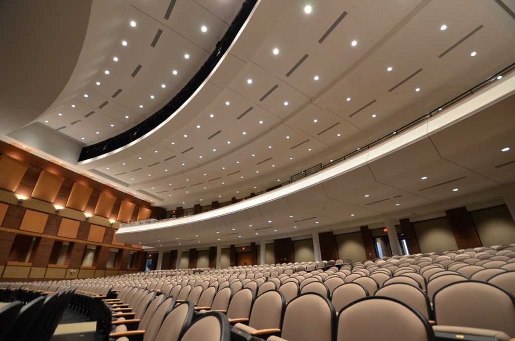 Empty beige seats arranged in rows inside a modern auditorium, illuminated by recessed lighting, with a curved ceiling, balcony area, and warm wood accents throughout the space.