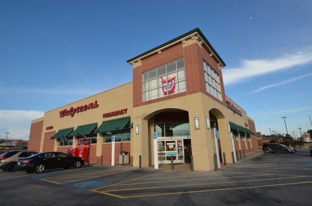 A Walgreens pharmacy storefront with cars parked in front, green awnings on the windows, and a clear sky above. Handicapped parking spaces are visible close to the entrance.