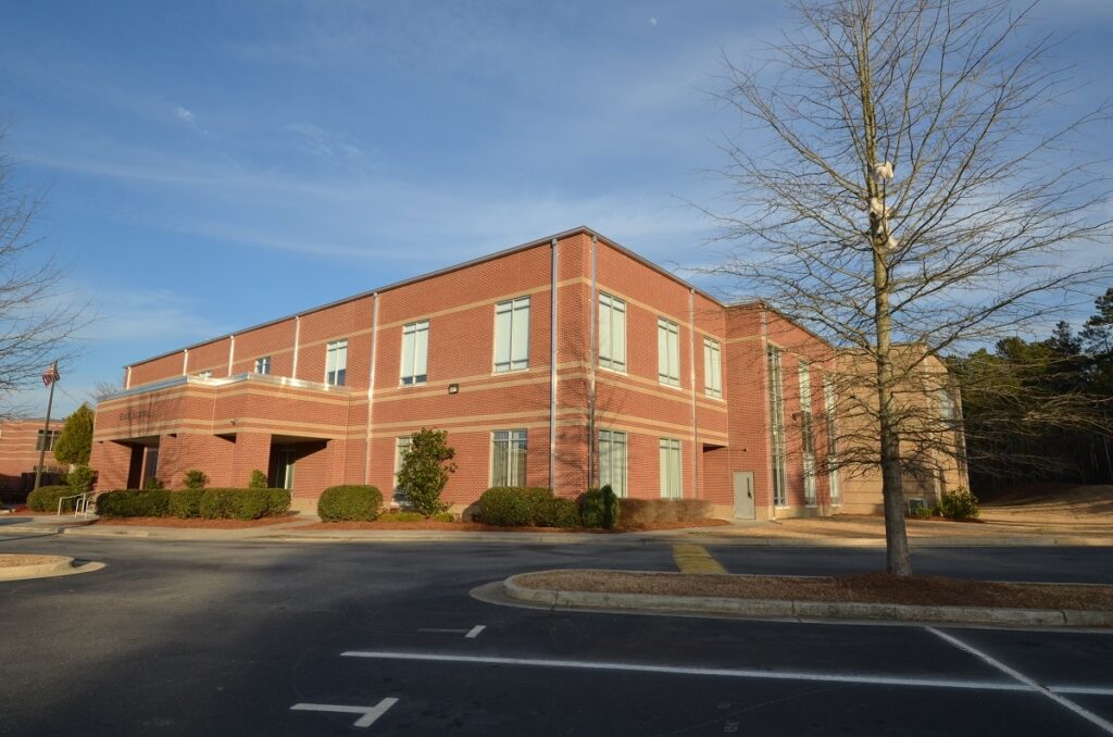 A two-story red brick building with numerous windows stands amid small bushes and trees, centered in an empty parking lot beneath a clear blue sky.