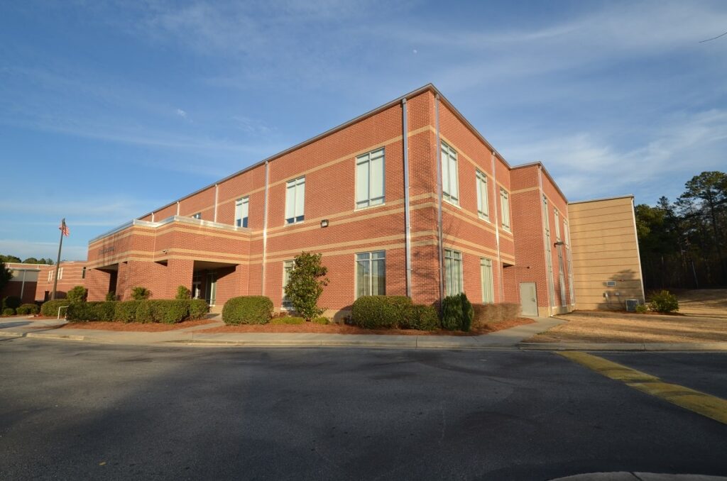 A two-story brick building featuring numerous windows stands amid bushes and trees beneath a clear sky. A paved driveway runs beside the building, bordered by a grassy area.